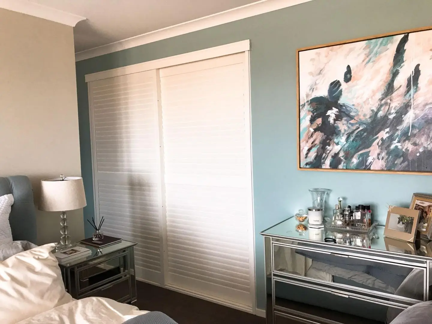 A bedroom features a mirrored dresser, abstract art, and a white wardrobe.