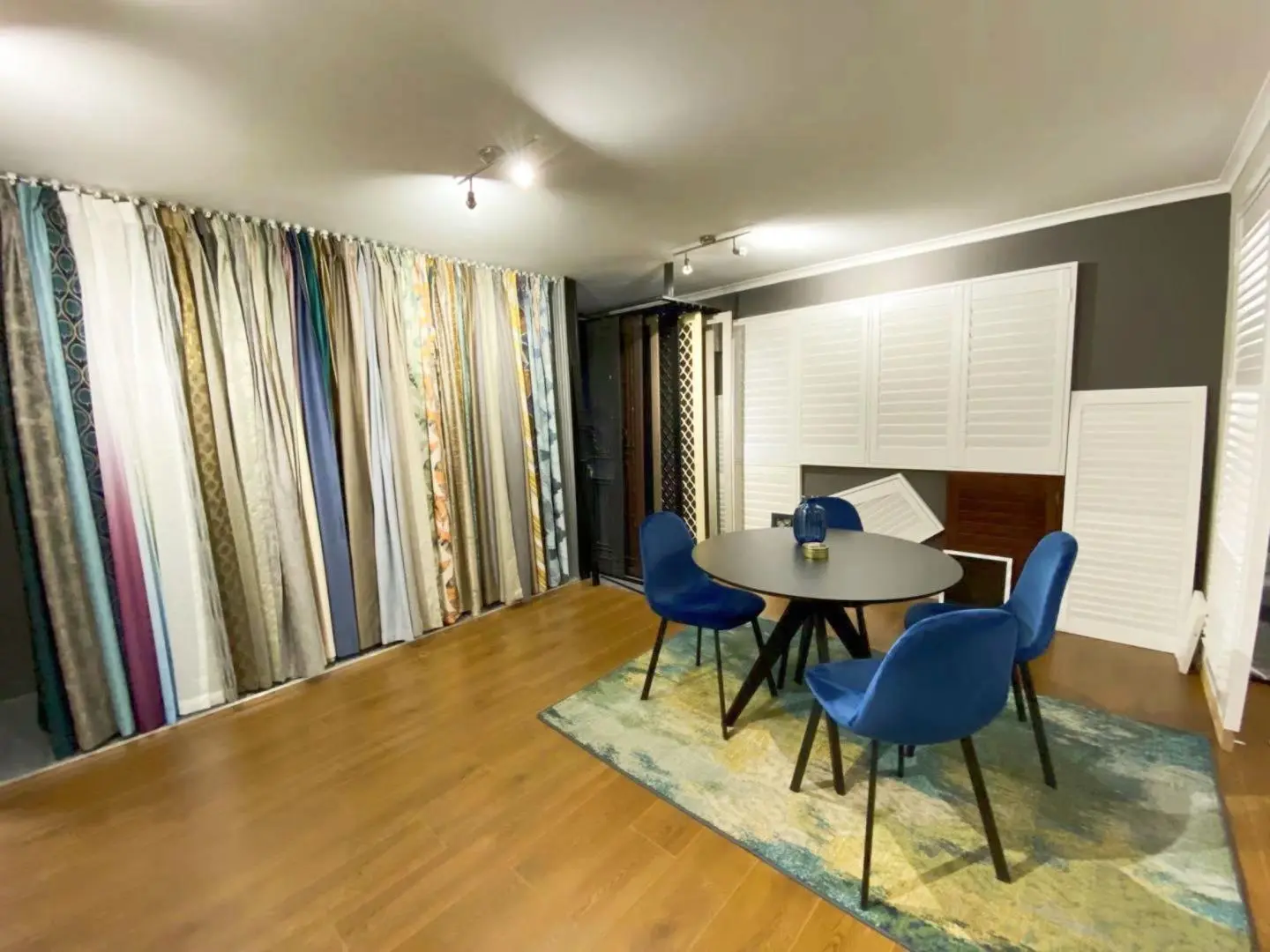 Curtains and shutters are displayed in a showroom with blue chairs and a table.
