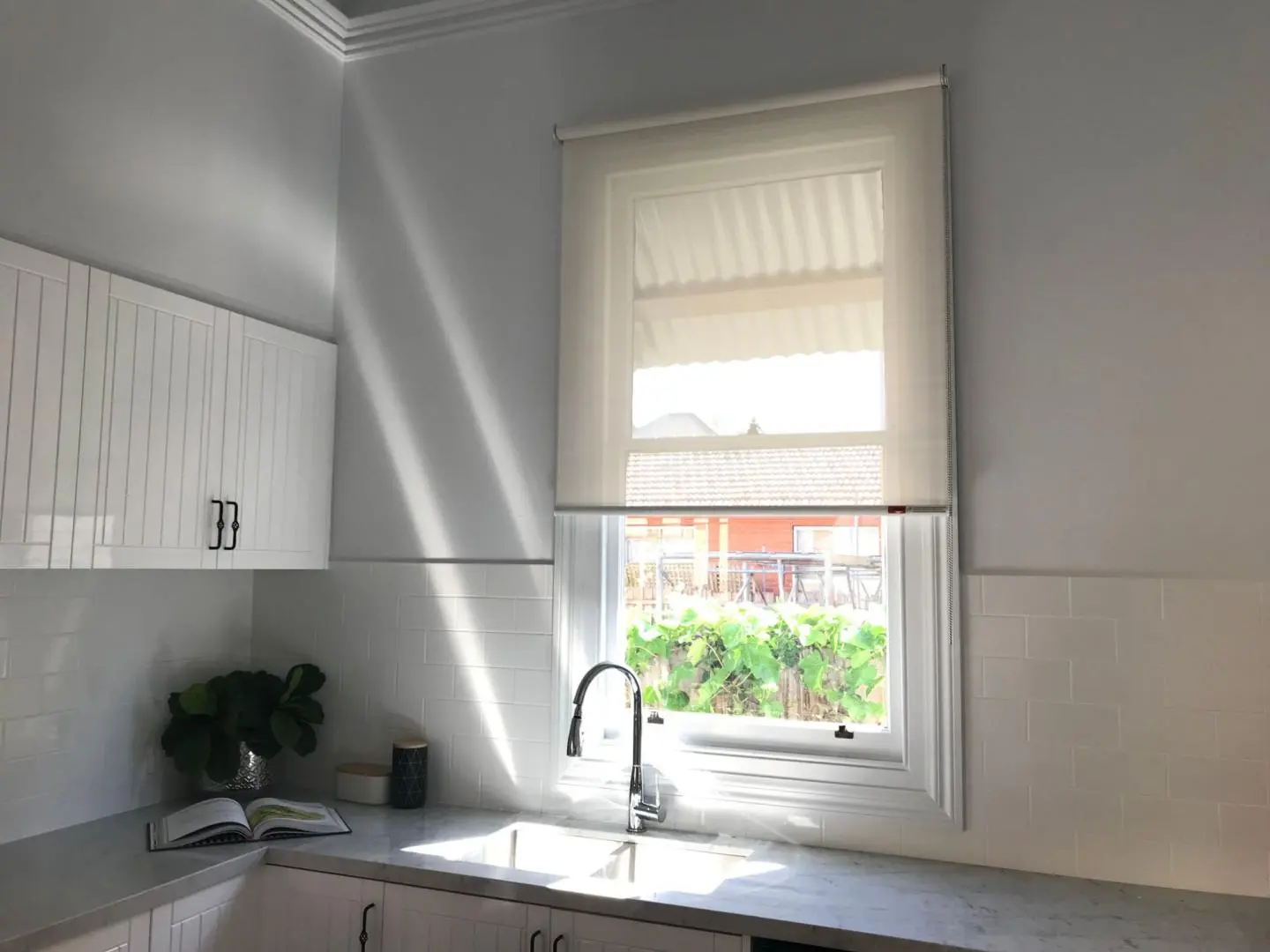 Sunlight streams into a modern kitchen with white cabinetry, subway tiles, and sheer roller blinds.