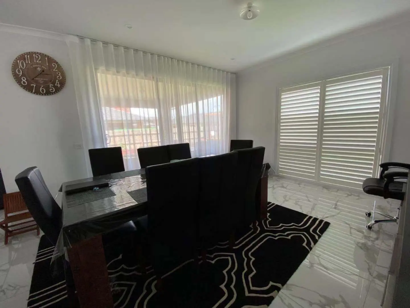A modern dining room features white shutters, a stylish rug, and a large clock on the wall.