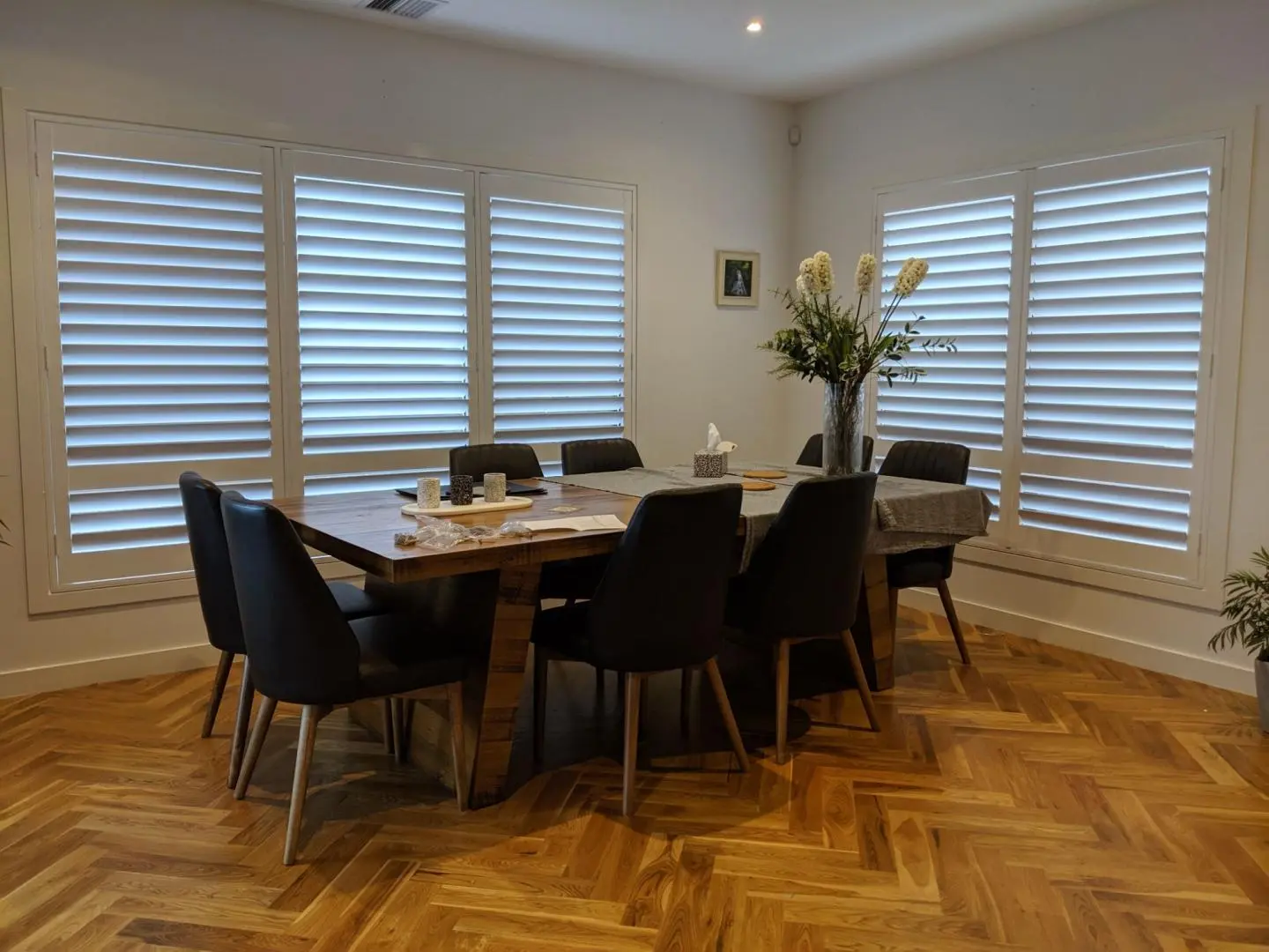 A modern dining room features a wooden table, black chairs, and stylish white shutters.