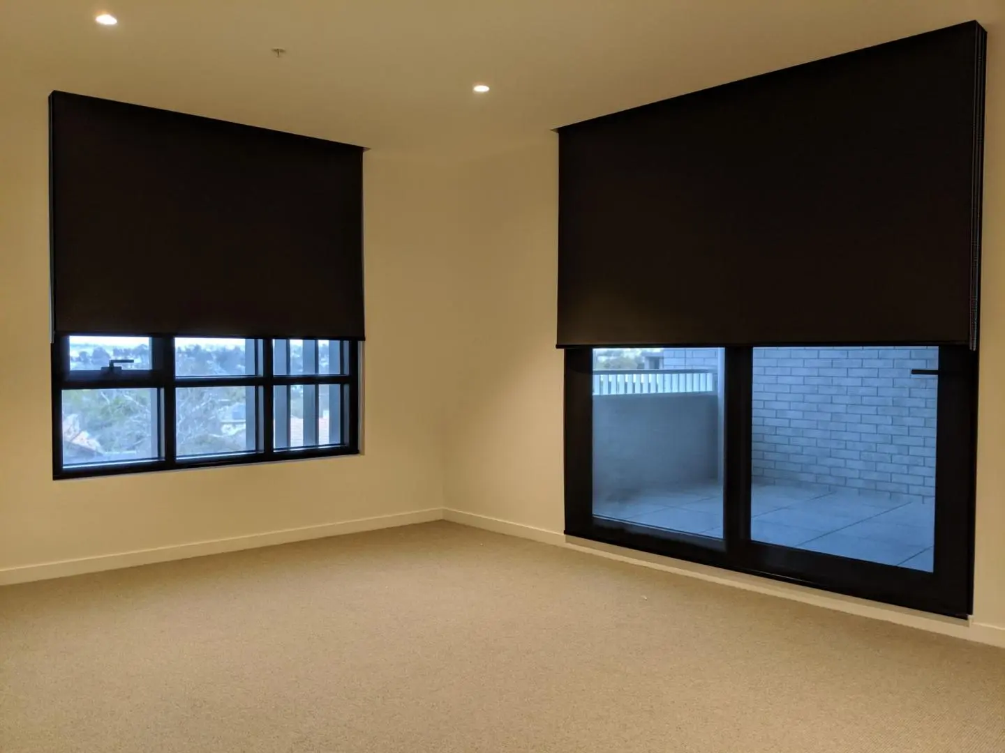 Empty modern room features dark roller blinds on a large window and a glass door.