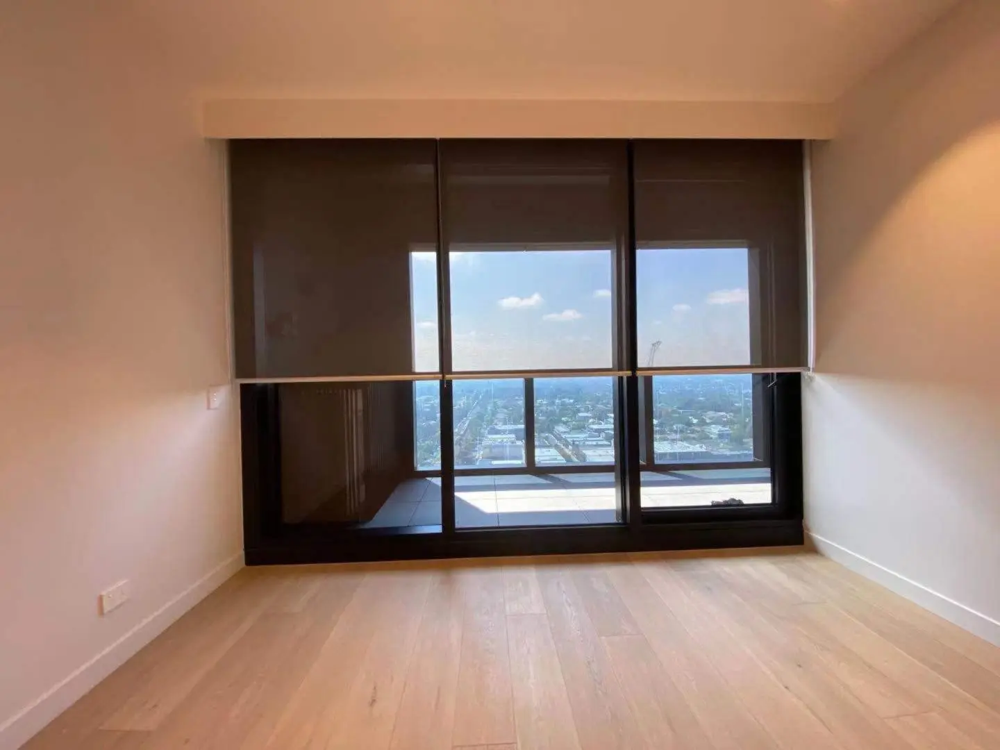 Empty room with hardwood floors, a large window, and dark roller blinds with a city view.