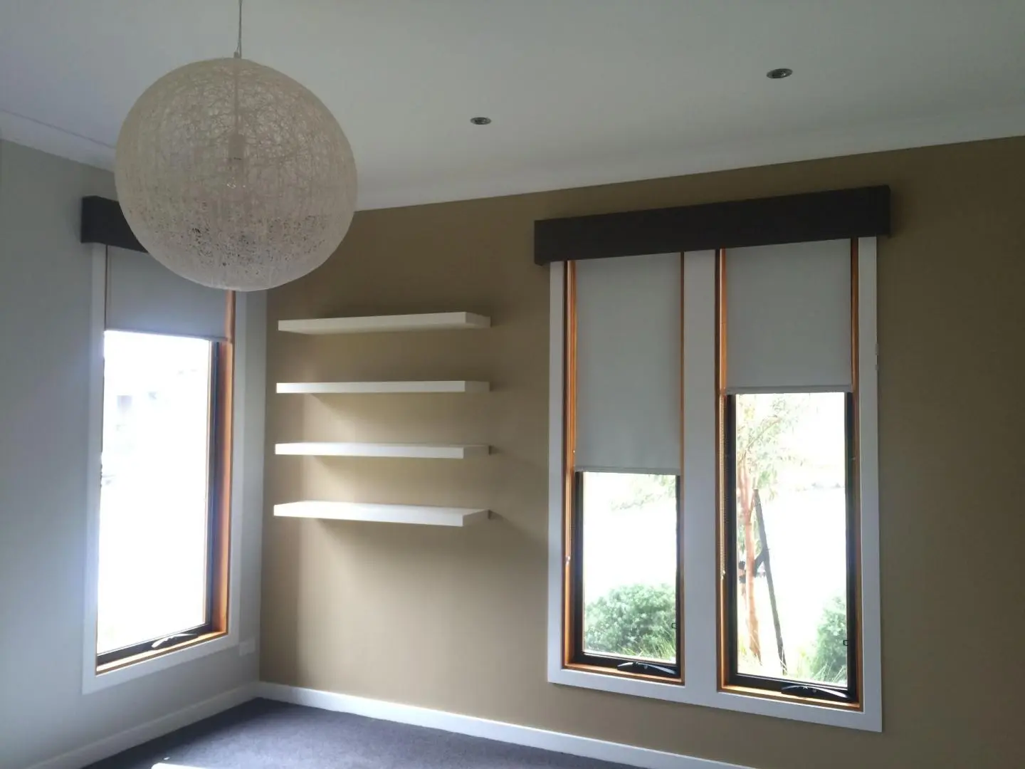 Bright modern empty room with large windows, light-filtering roller blinds, white floating shelves, and a spherical pendant light.