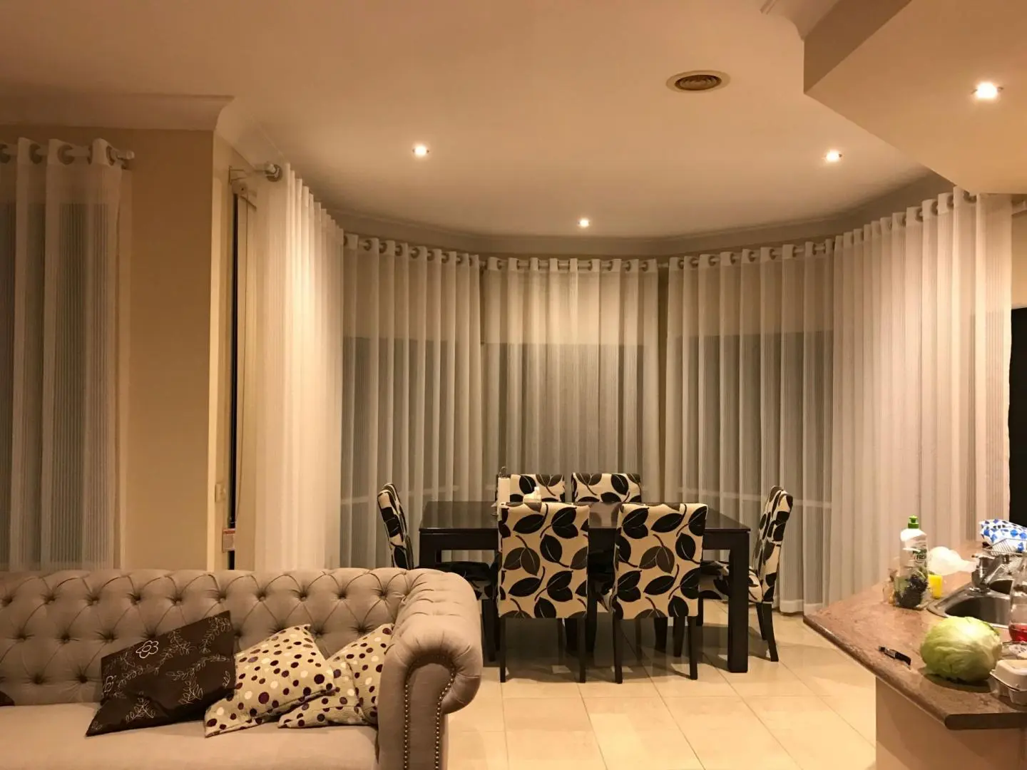 A well-lit living room features sheer curtains, a dining table with chairs, and a comfortable sofa.