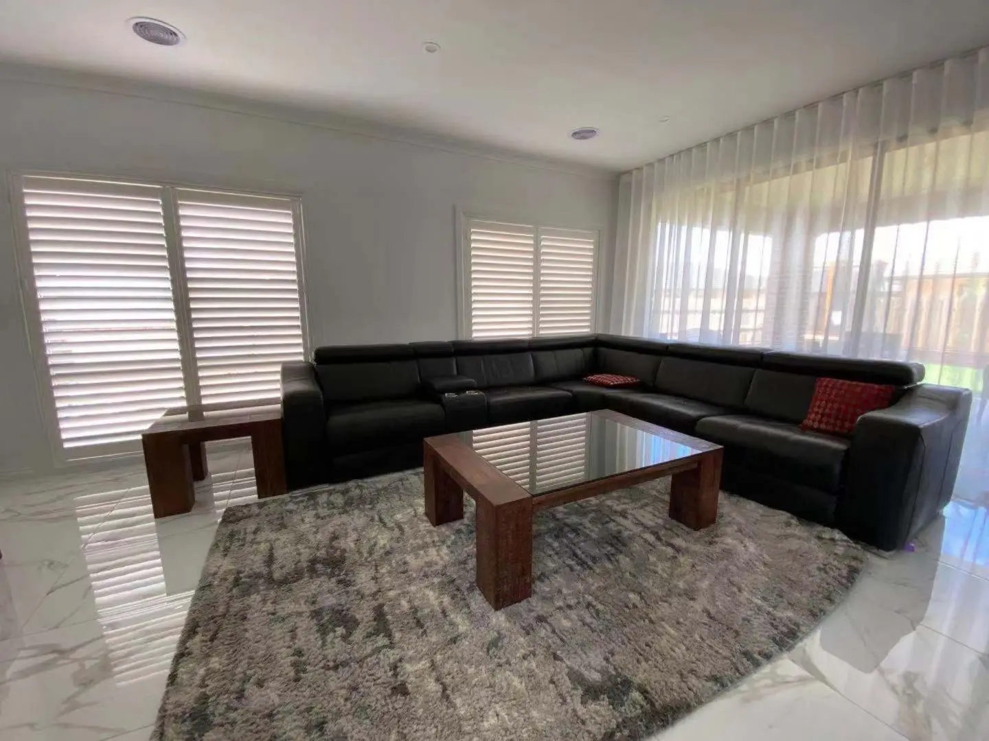 A modern living room features a black sectional sofa, rug, and glass-top coffee table, with window shutters and sheer curtains.