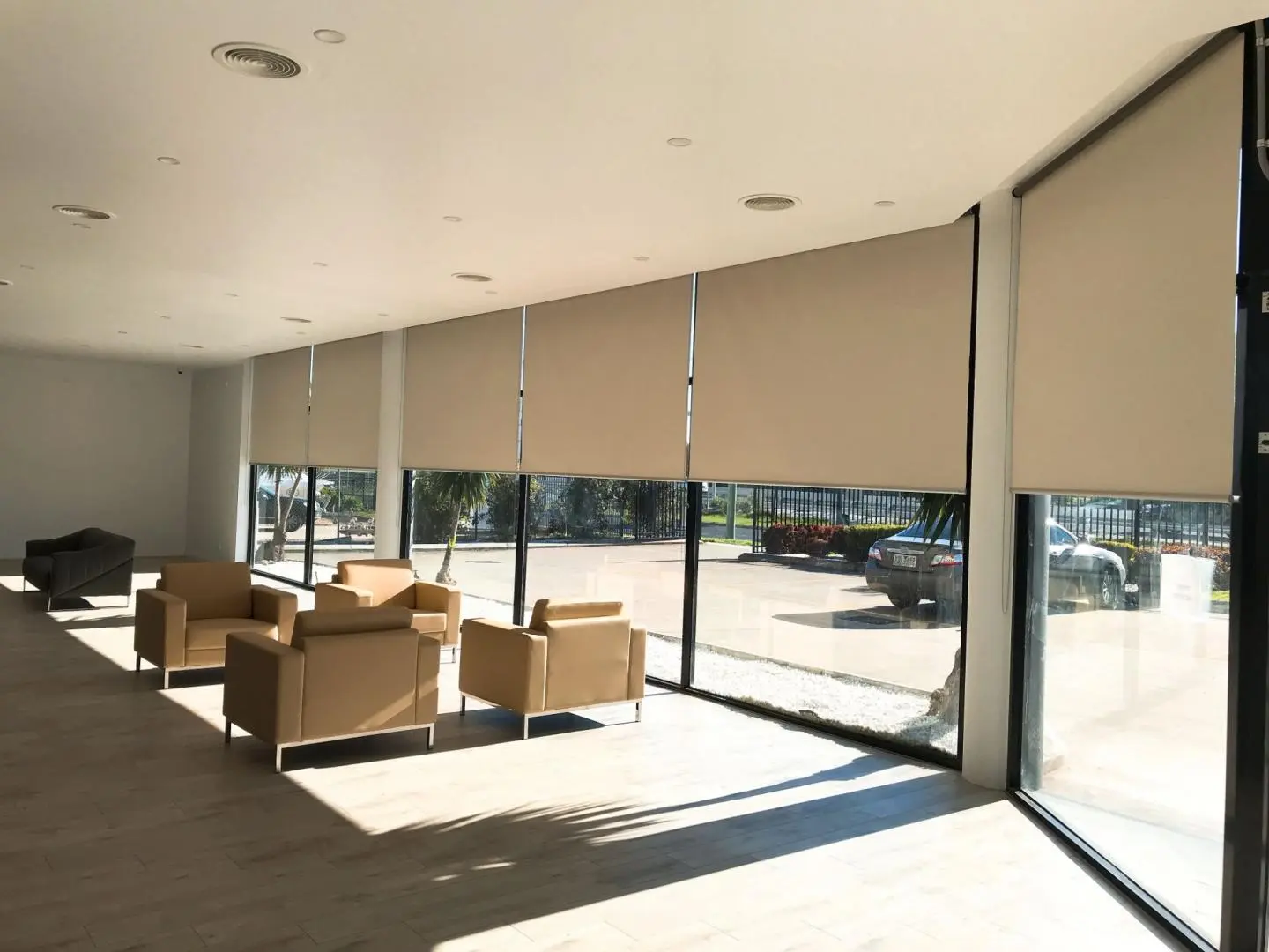 A modern lounge with large windows, beige roller blinds, and comfortable armchairs, brightened by natural light.
