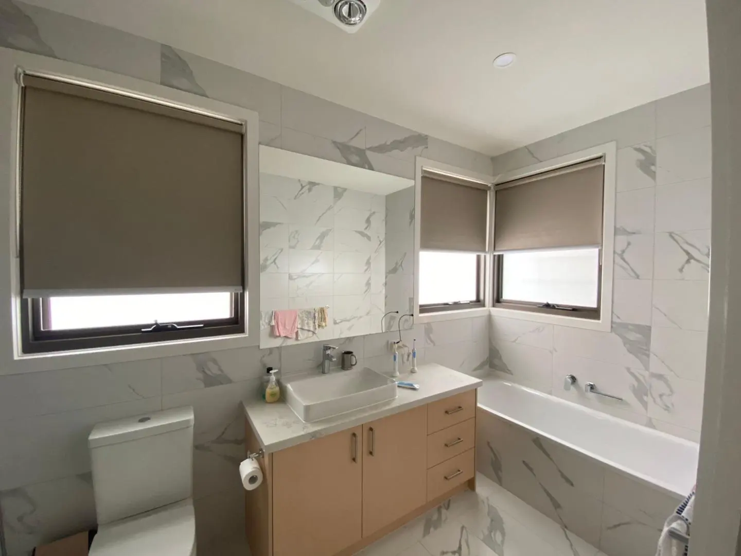 Modern bathroom with marble-look tiles, a light wood vanity, bathtub, toilet, and roller blinds.