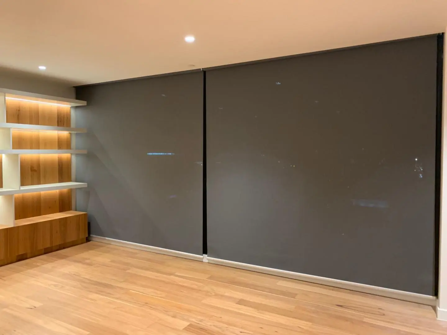 Modern room with dark roller blinds and illuminated built-in wooden shelves.