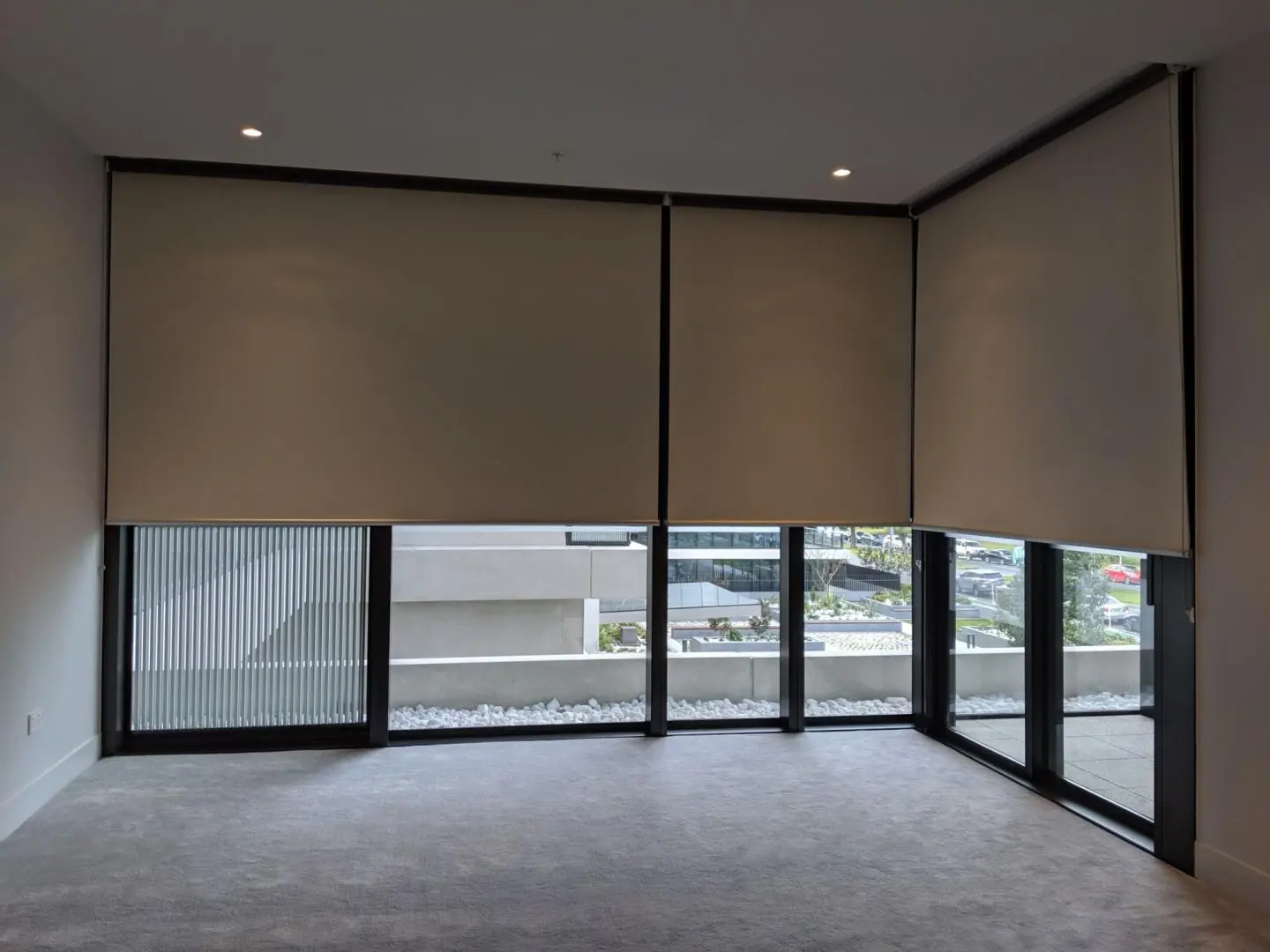 Beige roller blinds are partially lowered on large windows in a room, offering an outdoor view.