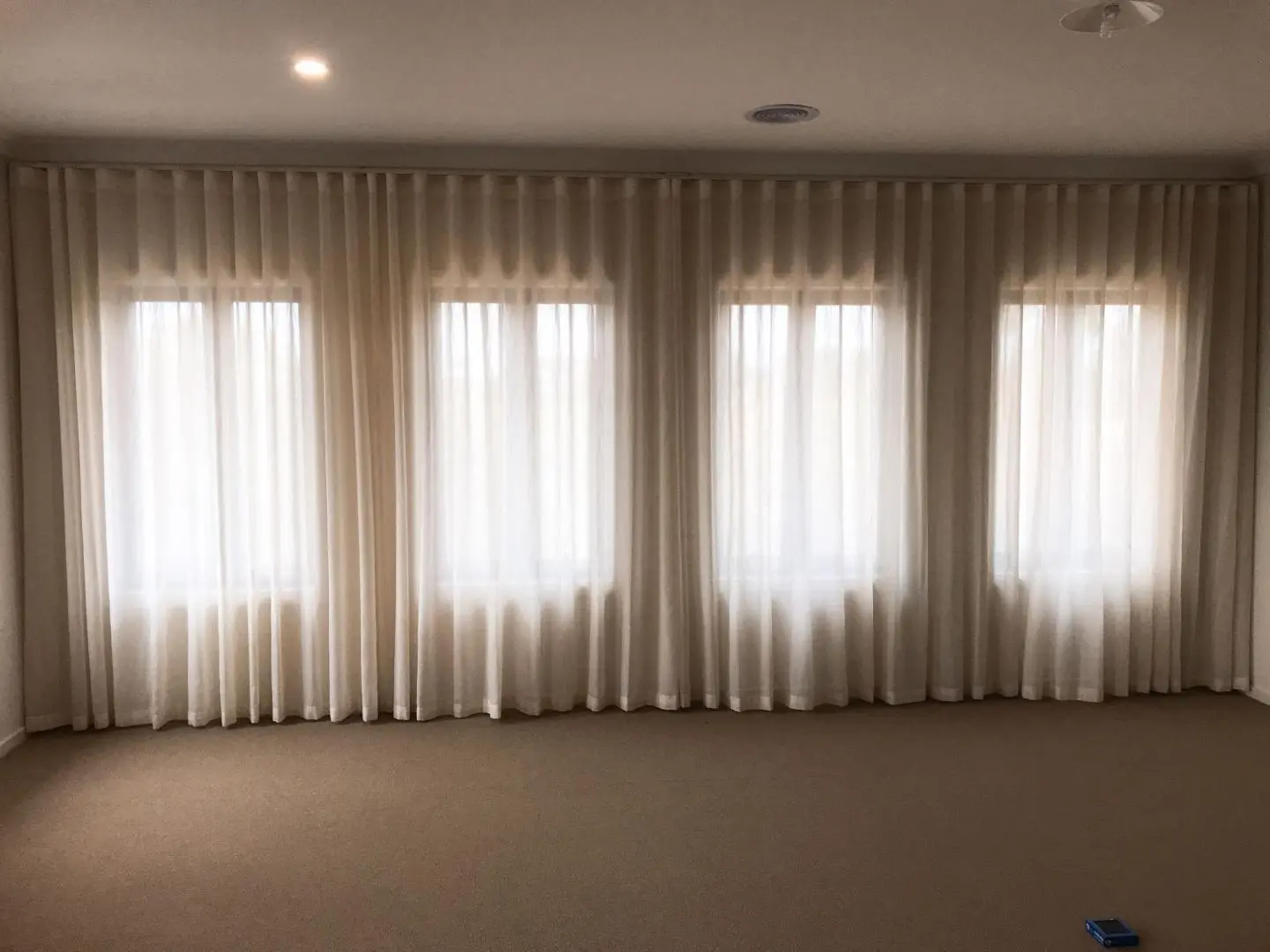 Bright, empty room features floor-to-ceiling sheer curtains on four windows and a light beige carpet.