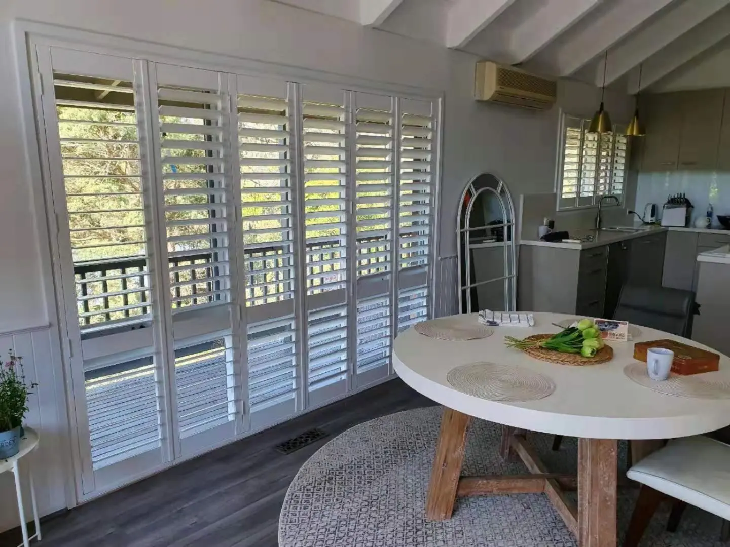 Bright living space with white shutters, a round table, and a kitchen in the background.