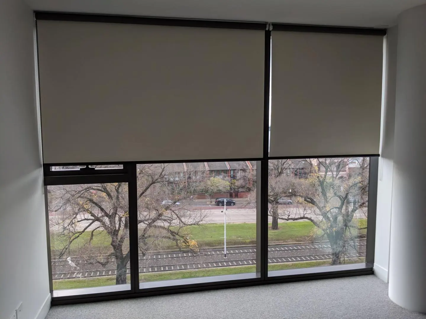 Modern roller blinds are partially lowered on large windows, offering an urban view with trees and train tracks.