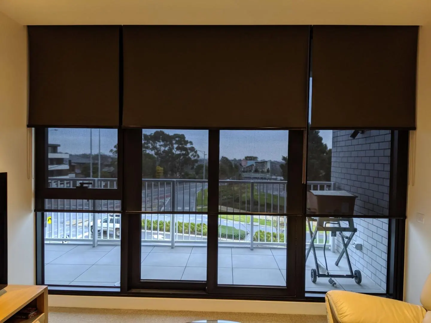 Dark roller blinds on large living room windows frame a balcony with a grill and urban landscape beyond.